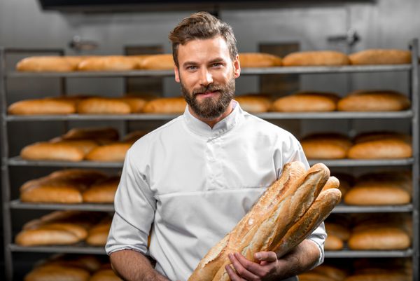 نانوایی خوش تیپ با لباس یکنواخت نان باگت با قفسه های نان در پس زمینه در محل تولید