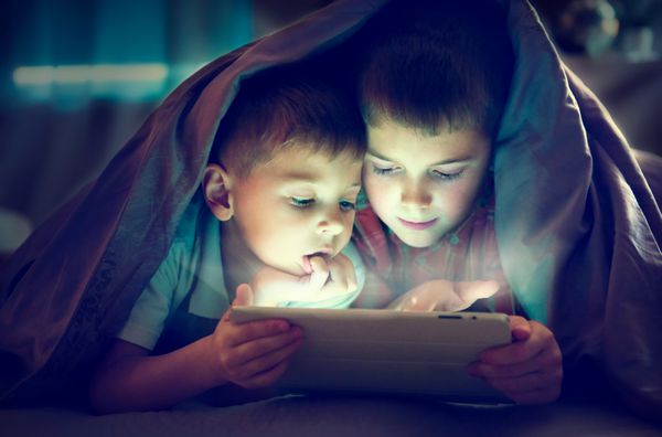 دو کودک شبانه از رایانه لوحی زیر پتو استفاده می کنند برادران با رایانه لوحی در یک اتاق تاریک