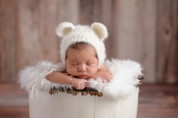یک نوزاد دختر یازده روزه که در یک سطل چوبی کوچک می خوابد او یک کلاه قلاب بافی و سفید خرس به سر دارد در استودیو روی پس‌زمینه چوبی