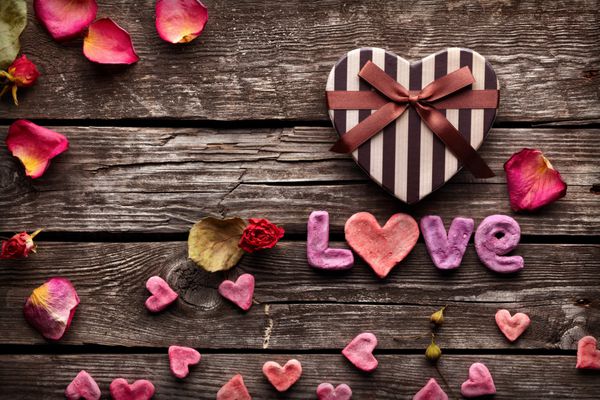 کلمه عشق با جعبه هدیه روز به شکل قلب روی بشقاب های چوبی قدیمی پس زمینه شیرین تعطیلات با گلبرگ های رز و قلب های کوچک