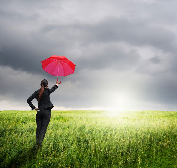 زن چتر تجاری با چتر قرمز در کنار ابرهای باران در علفزار ایستاده است
