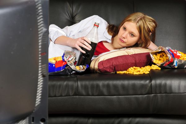 زن جوانی نوجوانی با موهای بلند روی مبل چرمی مشکی می‌چرخد در حال تماشای تلویزیون و خوردن چیپس و کوکاکولا