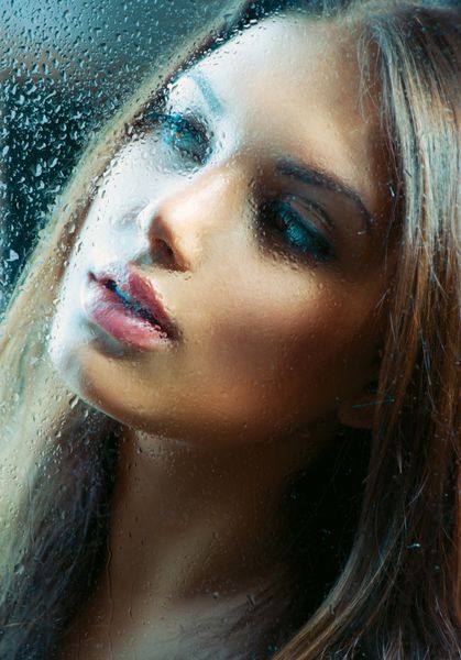 پرتره دختر زیبایی پشت شیشه خیس زن مالیخولیایی باران مدل زیبا که از پنجره با قطره های باران نگاه می کند