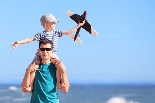 مرد جوان مثبت خندان و پسر هیجان زده اش که روی شانه های پدر و یک هواپیمای اسباب بازی در ساحل نشسته اند