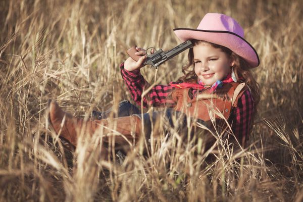 کودک ناز در بیرون از خانه تفریح ​​می کند دختر کوچک کابوی در مزرعه گندم هنگام غروب آفتاب دختر بچه خوشحال با تفنگ اسباب بازی و کلاه کابوی از طبیعت لذت می برد دختر گاوی آمریکایی پرتره کودک نوپا با لبخند زیبا