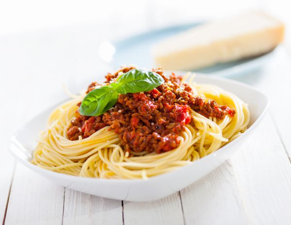 بشقاب سالم اسپاگتی ایتالیایی که روی آن گوجه فرنگی خوشمزه و سس بولونیز گوشت چرخ کرده و ریحان تازه روی میز چوبی سفید روستایی قرار گرفته است