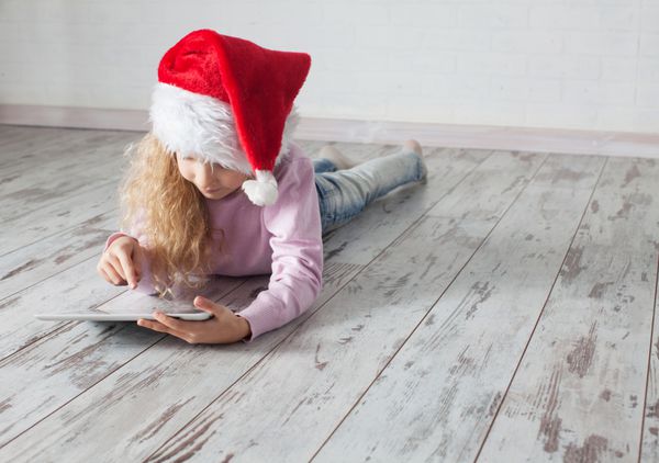 کودک با کلاه کریسمس با تبلت روی زمین خوابیده است کامپیوتر دختر