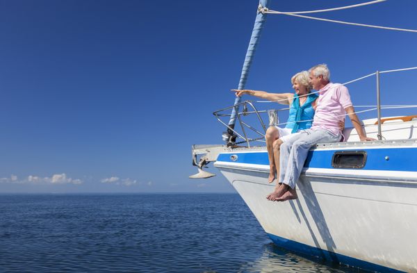 یک زوج سالمند شاد که در کنار یک قایق بادبانی در دریای آبی آرام نشسته اند و به افقی روشن اشاره می کنند
