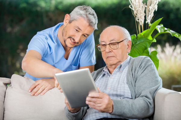 سرایدار مرد به مرد سالمند در استفاده از تبلت دیجیتال در ایوان خانه سالمندان کمک می کند