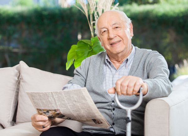 پرتره مرد سالخورده مطمئن با روزنامه و عصا نشسته روی مبل در ایوان خانه سالمندان