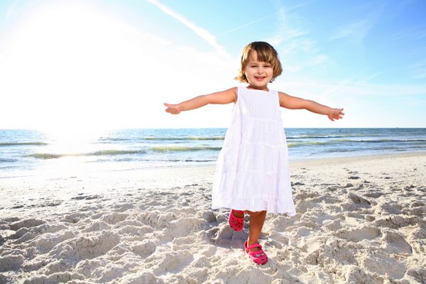 دختر کوچک شاد با لباس سفید در حال لذت بردن از روز آفتابی در ساحل در مقابل خورشید شلیک کنید