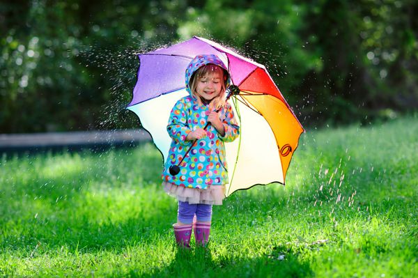 دختر نوپا ناز بامزه ای با پوشیدن کت ضد آب با چتر رنگارنگ در باغ در روز بارانی و آفتابی