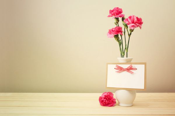 گل های میخک صورتی زیبا روی گلدان سفید با کارت تبریک خالی روی میز چوبی با پس زمینه دیوار قهوه ای روشن