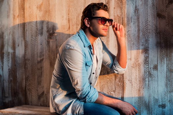 استراحت در زیر اشعه خورشید مرد جوان خوش تیپ به سمت دیوار نگاه می کند و عینک آفتابی خود را در حالی که روی دیوار نشسته است تنظیم می کند