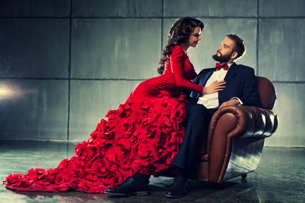 زوج عاشق جوان زیبا در پرتره لباس شب زن قرمزپوش و مردی با کت و شلوار مشکی که روی صندلی نشسته است