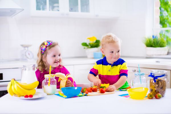 دختر و پسر کوچک در حال آماده کردن صبحانه در آشپزخانه غذای سالم برای کودکان کودک در حال خوردن شیر و خوردن میوه کودک شاد پیش دبستانی از وعده غذایی صبحگاهی غلات موز و توت فرنگی لذت می برد بچه ها در حال آشپزی