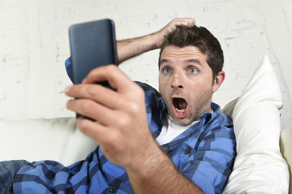 مرد جوان جذابی که روی مبل خانه دراز کشیده و با استفاده از اینترنت روی تلفن همراه خود در حال کشیدن مو در مفهوم استرس تکنولوژی شگفت زده و شوکه شده است