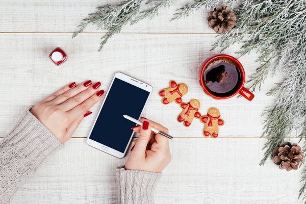 دست نوشته زن آراسته روی تلفن همراه کوکی‌های شیرینی زنجبیلی و فنجان چای کریسمس روی پس‌زمینه چوبی برفی پردازش به نگاه فیلم رترو