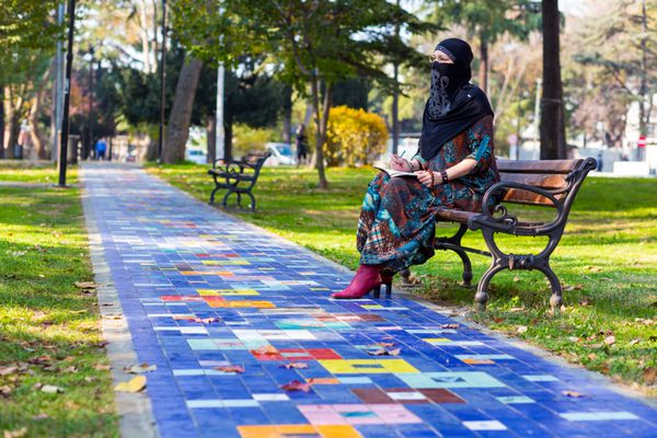 زن جوان اسلامی روی نیمکت پارک در امتداد کوچه سنگفرش رنگارنگ نشسته و در دفترچه یادداشت کاغذی یادداشت های دستی می نویسد