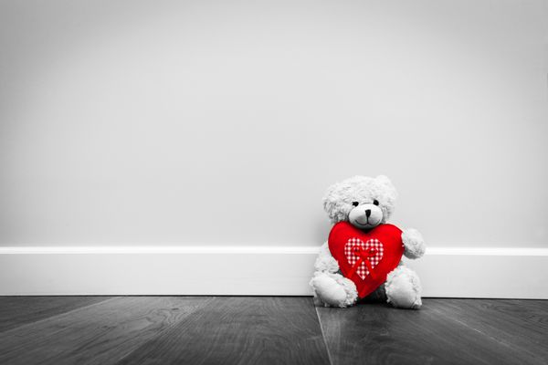خرس عروسکی زیبا با قلب بزرگ قرمز مخملی روی زمین چوبی روی دیوار سفید نشسته است سبک روستایی قدیمی روز عشق