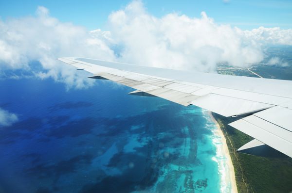 نمایی از پنجره هواپیما بال یک هواپیما که بر فراز ابرها بر فراز جزیره گرمسیری پرواز می کند