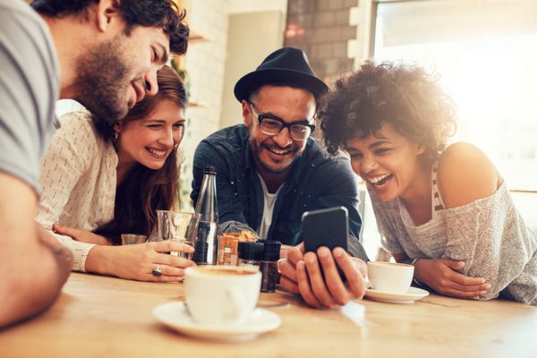 پرتره دوستان جوان شادی که در حالی که در کافه نشسته اند به تلفن هوشمند نگاه می کنند افراد مختلط با استفاده از تلفن همراه پشت میز رستوران نشسته اند