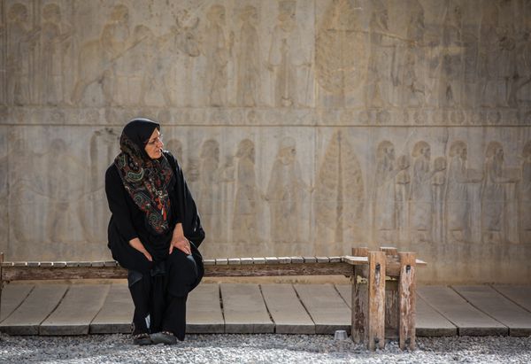 تخت جمشید ایران - حدود آوریل 2015 بانویی با لباس های سنتی روی نیمکتی مقابل دیوار حکاکی شده باستانی نشسته است
