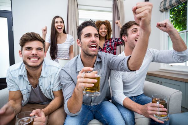 دوستانی که هنگام تماشای مسابقه فوتبال در تلویزیون تشویق می کنند و نوشیدنی می نوشند