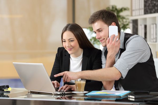 دو همکار که به صورت آنلاین با هم کار می کنند با یک لپ تاپ دستورالعمل می دهند و روی میز دفتر با تلفن تماس می گیرند