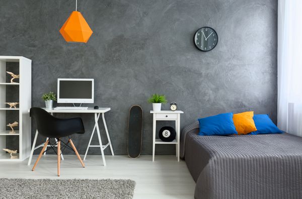 اتاق خواب با دیوارهای خاکستری و تخت خاکستری با بالش های رنگارنگ و لامپ نارنجی کنار تخته اسکیت دیواری و میز با کامپیوتر