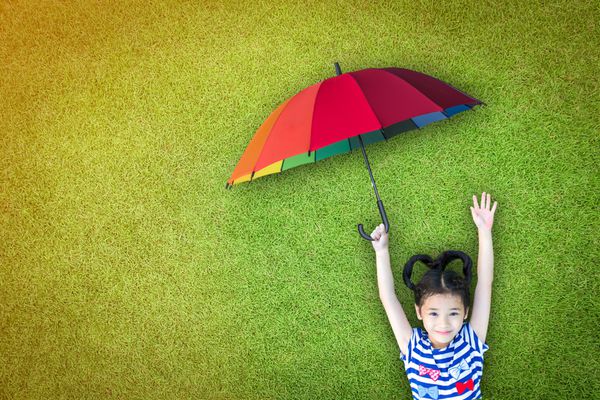 بچه دختر آسیایی شاد که دستش را بلند می کند و روی زمین چمن سبز چمن طبیعی دراز کشیده است و چتر رنگارنگ رنگین کمان محافظ UV را در روز آفتابی نگه می دارد مفهوم حفاظت از بیمه ایمنی مراقبت بهداشتی csr