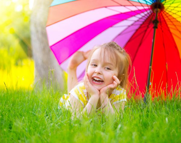 پرتره یک دختر کوچک خندان که روی چمن سبز زیر چتر رنگارنگ دراز کشیده است کودک ناز در حال لذت بردن از طبیعت در فضای باز بچه سالم و بی خیال بیرون در پارک تابستانی محافظت در برابر آفتاب
