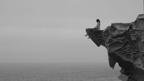 دختری تنها در لبه صخره در اقیانوس مه نشسته و به دنبال چیزی با رنگ سیاه و سفید است