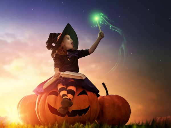 هالووین مبارک جادوگر کوچولوی شاد و بامزه با عصای جادویی و کتاب طلسم دختر بچه زیبا با لباس جادوگر که روی کدو تنبل بزرگ نشسته و تداعی می کند و می خندد