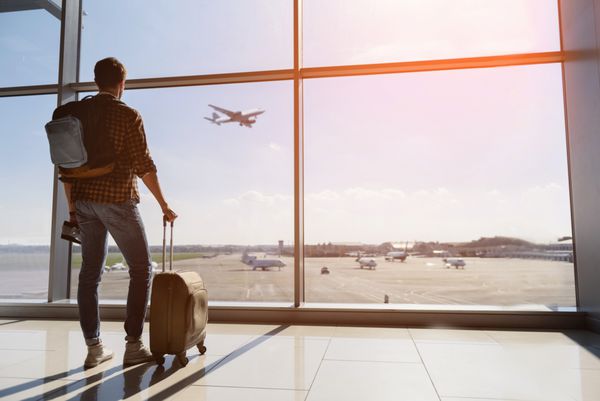 گردشگر مرد آرام در فرودگاه ایستاده و از پنجره به پرواز هواپیما نگاه می کند بلیت و چمدان در دست دارد غروب آفتاب