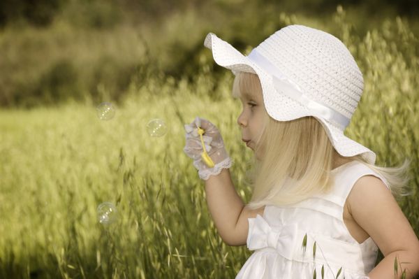 دختر کوچکی با لباس سفید و کلاهی که حباب‌های صابون را در یک مزرعه باد می‌کشد