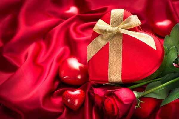 پس زمینه هدیه جعبه هدیه به شکل قلب قرمز و گل رز قرمز در زمینه ابریشم قرمز طراحی هنر مرزی