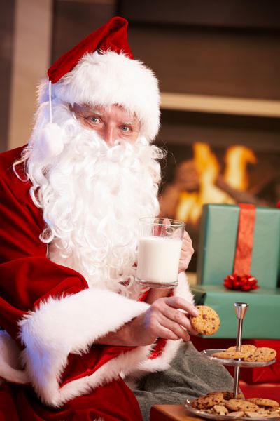 بابا نوئل راضی نشسته کنار firepl در حال خوردن شیر و خوردن شیرینی های شکلاتی است و به دوربین نگاه می کند