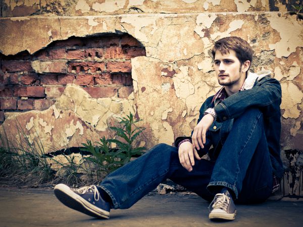 مرد جوانی که لباس شلوار جین پوشیده است روی زمین در مقابل دیوار ویران شده ترک خورده نشسته است