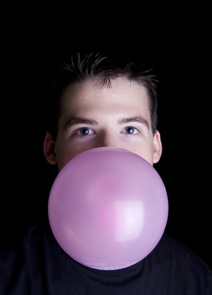 مرد جوان قفقازی با موهای قهوه ای در حال دمیدن حباب صورتی با آدامس حباب دار جدا شده روی پس زمینه سیاه
