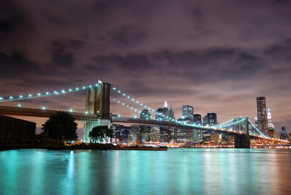نمای پانورامای شهر نیویورک منهتن با پل بروکلین در شب با آسمان‌خراش‌های ساختمان اداری که بر فراز رودخانه هادسون روشن شده است