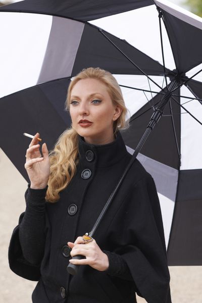 مدل زیبای ایستاده زیر چتر سیاه و سفید شیک در حال کشیدن سیگار