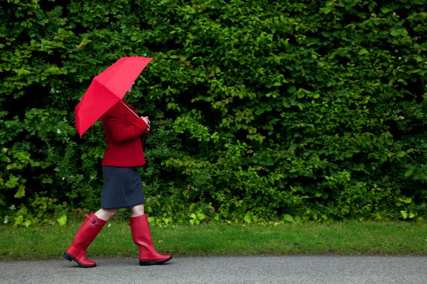 عکس زنی قرمزپوش که در یک روز ابری با شروع بارندگی در امتداد جاده ای با چترش بالا رفته است تاری حرکت خفیف روی پاهای او