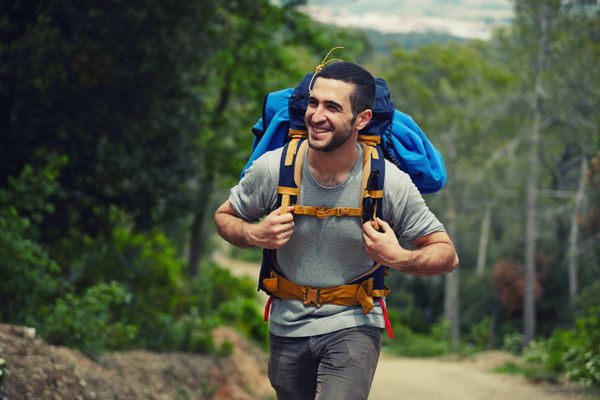 کوهنورد جوان با کوله پشتی که در مسیر راه می رود و لبخند می زند