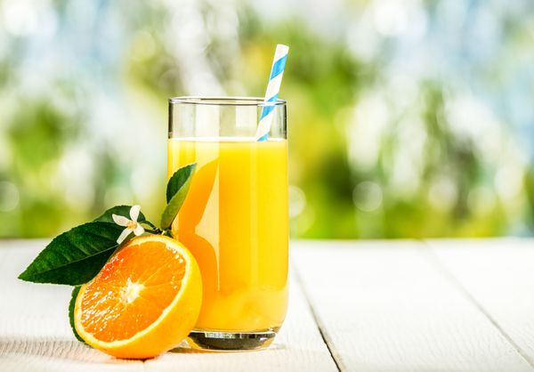لیوان بلند آب پرتقال تازه فشرده و خوشمزه که روی میز پیک نیک چوبی در فضای باز در یک روز تابستانی با یک پرتقال تازه نصف شده ایستاده است
