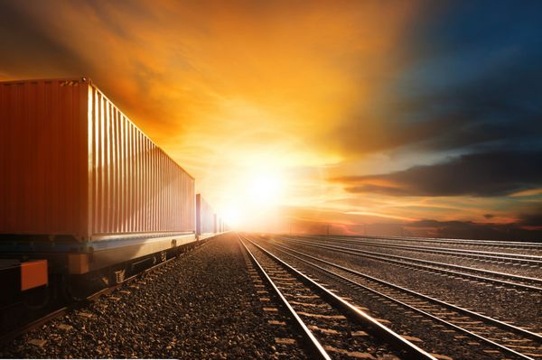 قطارهای کانتینری صنعتی که در مسیر راه آهن در برابر آسمان زیبای غروب خورشید حرکت می کنند برای حمل و نقل زمینی و تجارت لجستیک استفاده می کنند
