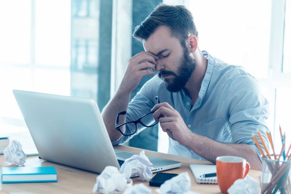 احساس خستگی مرد جوان ناامید ریش در حالی که در محل کار خود در دفتر نشسته است بینی خود را ماساژ می دهد و چشمانش را بسته نگه می دارد