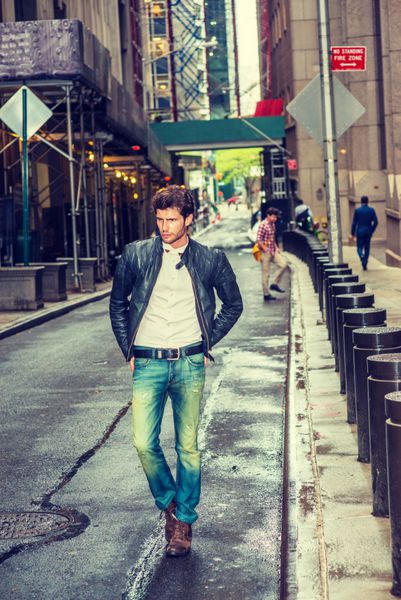 تاجر اروپایی در حال سفر در نیویورک کت چرمی مشکی پوشیده پیراهن زیر سفید شلوار جین آبی کفش چکمه قهوه ای دست روی باسن مردی با ریش که بعد از باران در خیابان مرطوب باریک راه می رود