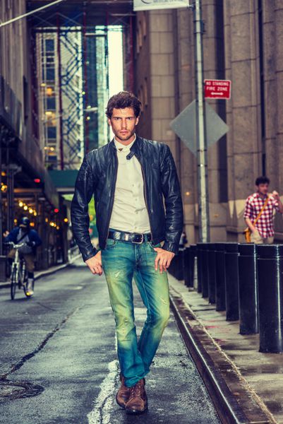تاجر اروپایی در حال سفر در نیویورک کت چرمی مشکی زیرپیراهن سفید شلوار جین آبی کفش چکمه قهوه‌ای پسر جوانی با ریش که بعد از باران در خیابان مرطوب باریک راه می‌رود