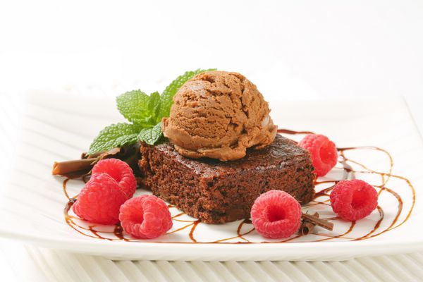 نمای نزدیک از کیک براونی با قاشق بستنی شکلاتی و تمشک روی بشقاب سفید و مات